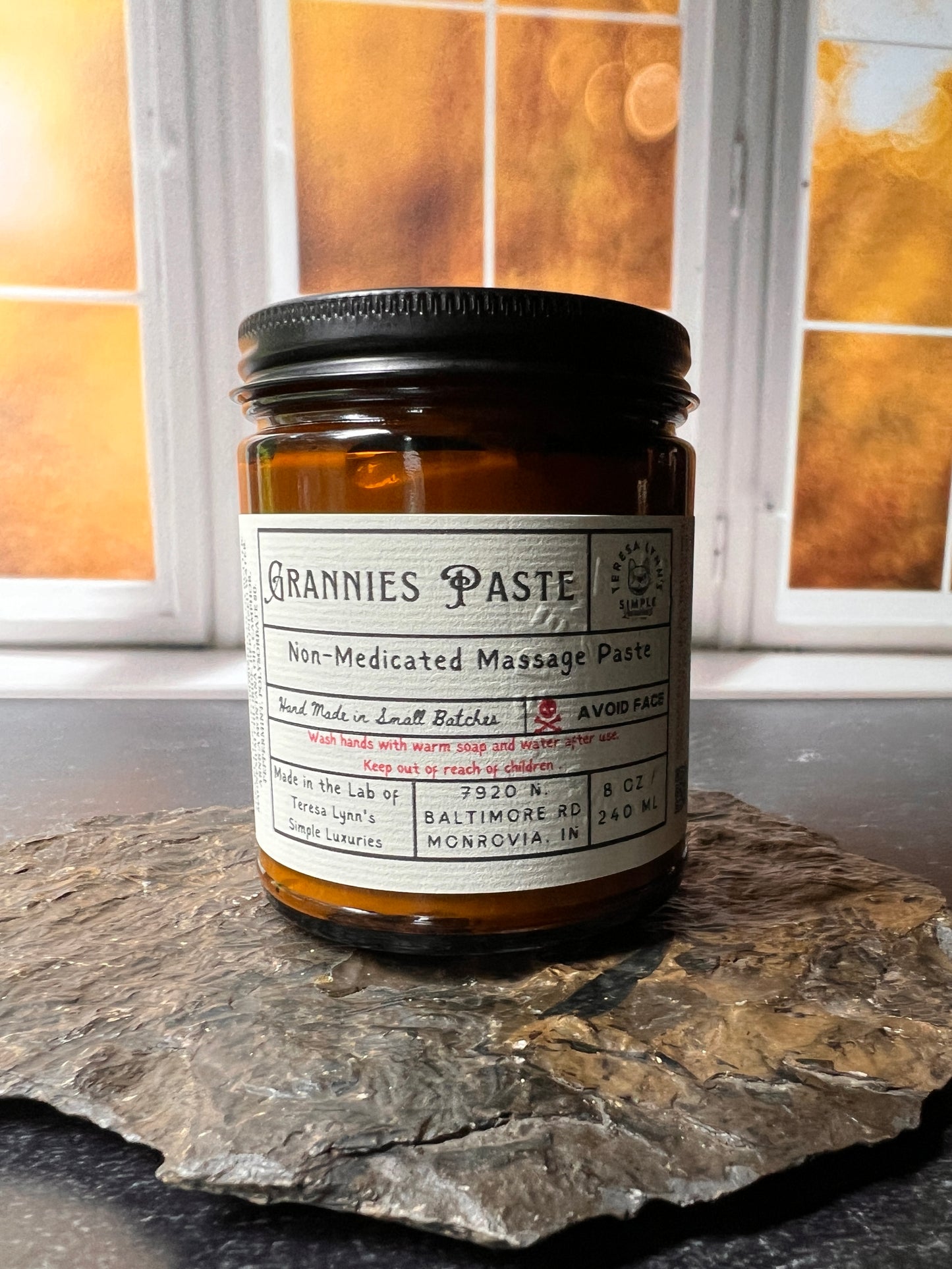 An Amber glass jar labeled Grannie Paste in a prescription inspired label. The jar has a black metal lid and an apothecary vibe. The background is aged metal. Her hand and the jar are the focus.