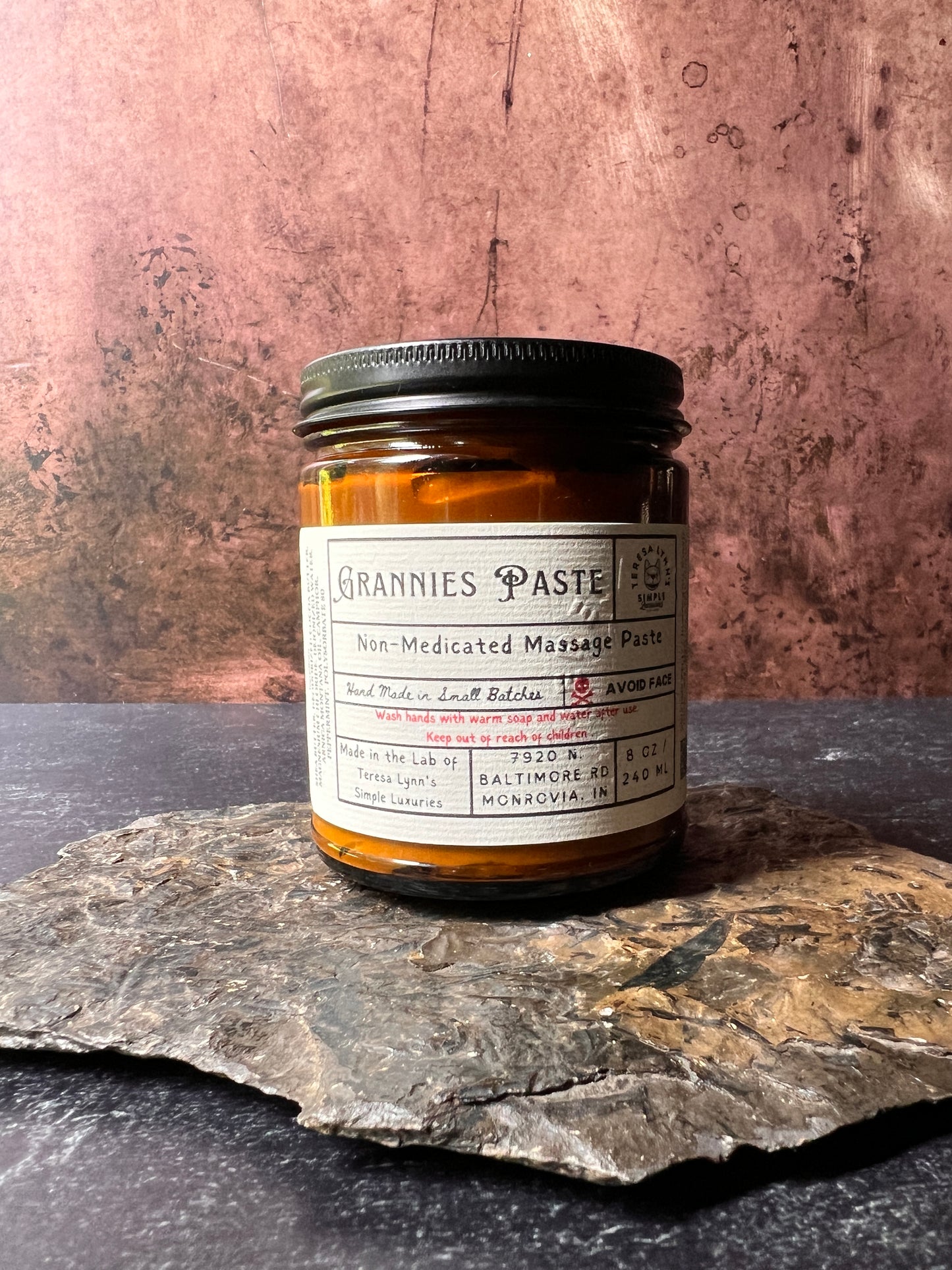An Amber glass jar labeled Grannie Paste in a prescription inspired label. The jar has a black metal lid and an apothecary vibe. The background is aged metal. Her hand and the jar are the focus. Nostalgic Grannies Paste - A Scent of Childhood- Herbal Massage Cream - The Preachers Daughter Candle Co