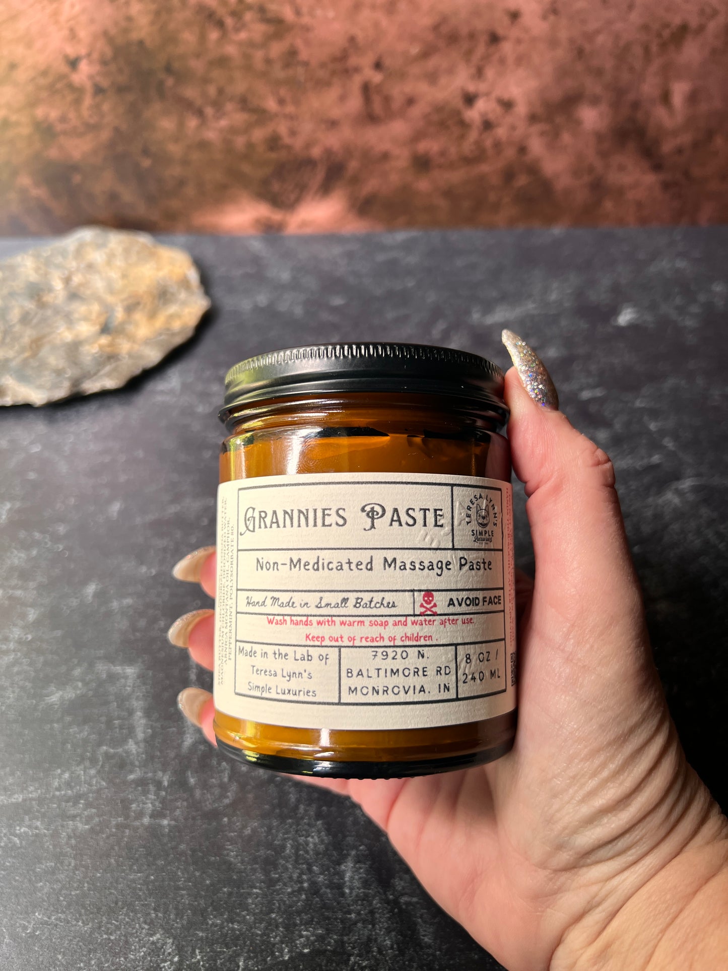 An Amber glass jar labeled Grannie Paste in a prescription inspired label. The jar has a black metal lid and an apothecary vibe. The background is aged metal. Her hand and the jar are the focus. Nostalgic Grannies Paste - A Scent of Childhood- Herbal Massage Cream - The Preachers Daughter Candle Co