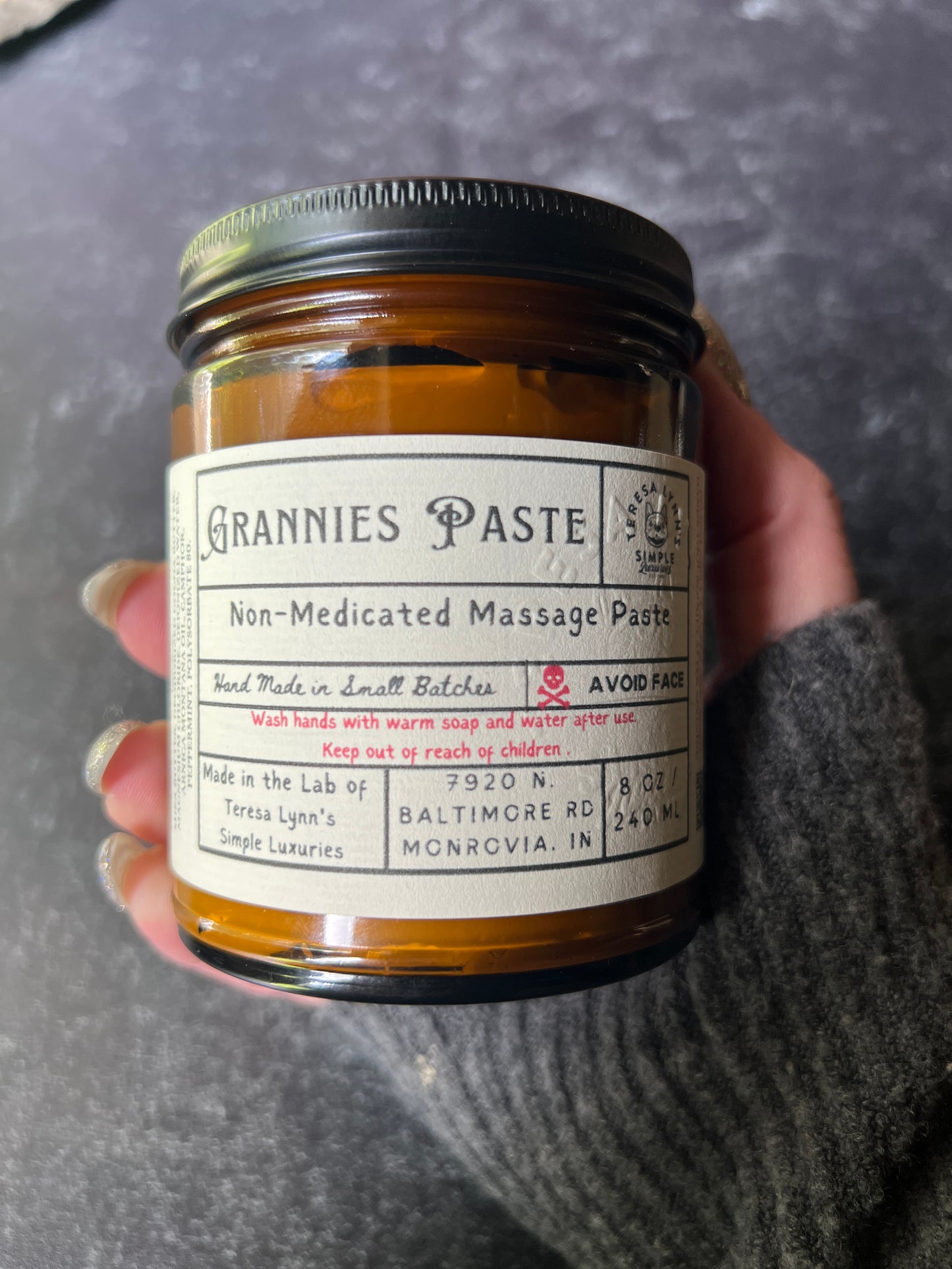 Teresa holds a brown jar in her hand labeled Grannie Paste in a prescription inspired label. The jar has a black metal lid and an apothecary vibe. The background is aged metal. Her hand and the jar are the focus.