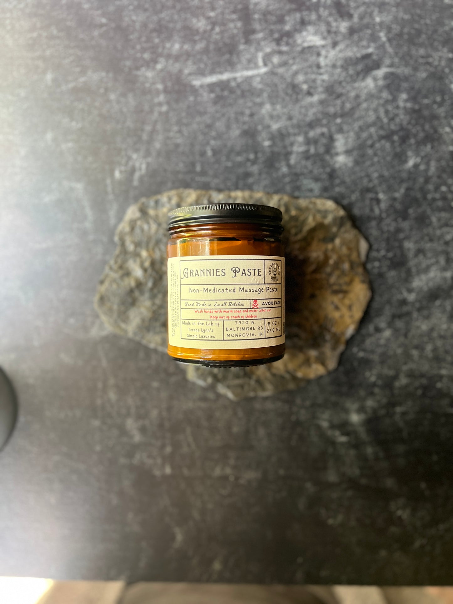 Teresa holds a brown jar in her hand labeled Grannie Paste in a prescription inspired label. The jar has a black metal lid and an apothecary vibe. The background is aged metal. Her hand and the jar are the focus.