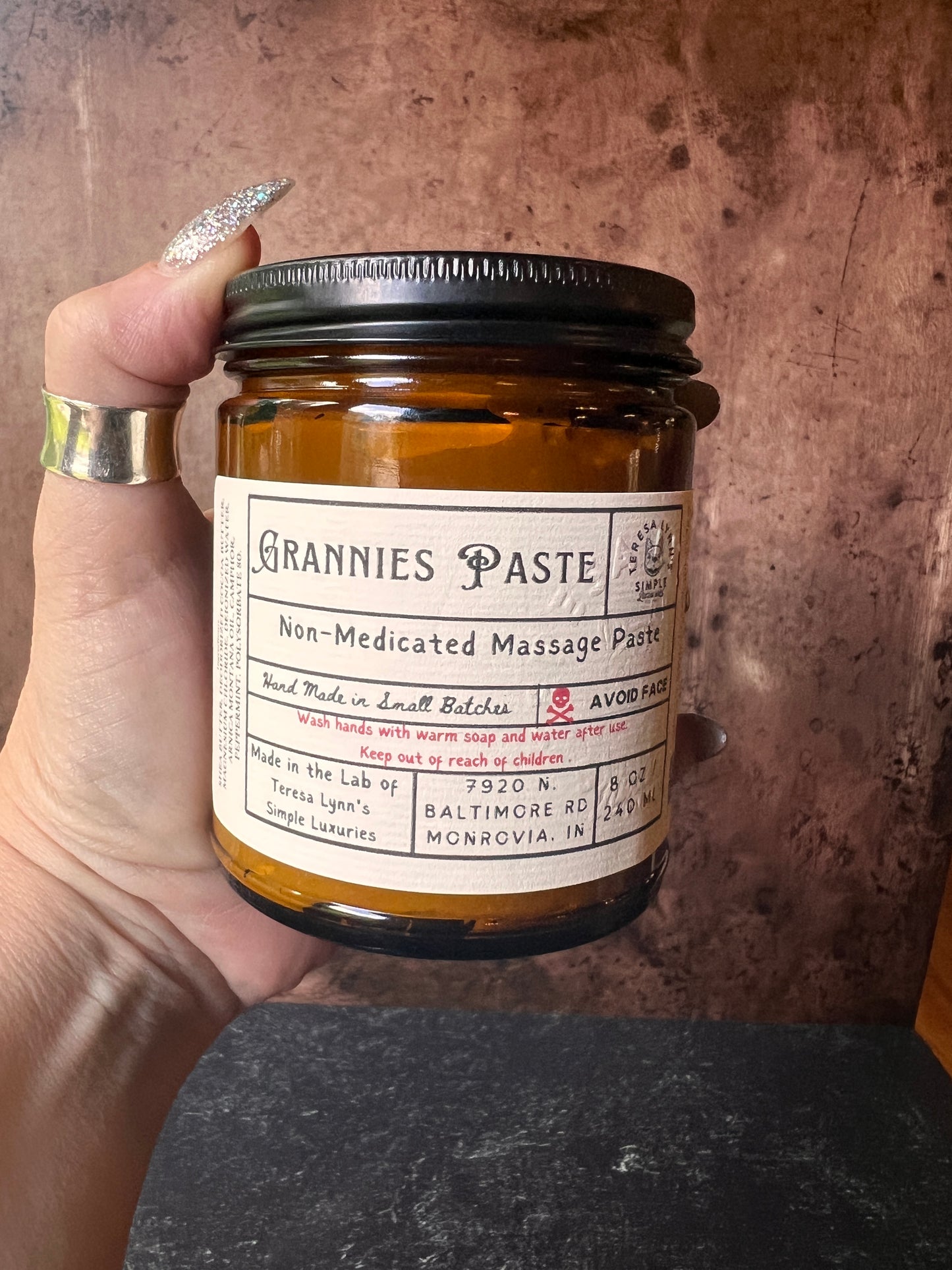 Teresa holds a brown jar in her hand labeled Grannie Paste in a prescription inspired label. The jar has a black metal lid and an apothecary vibe. The background is aged metal. Her hand and the jar are the focus.
