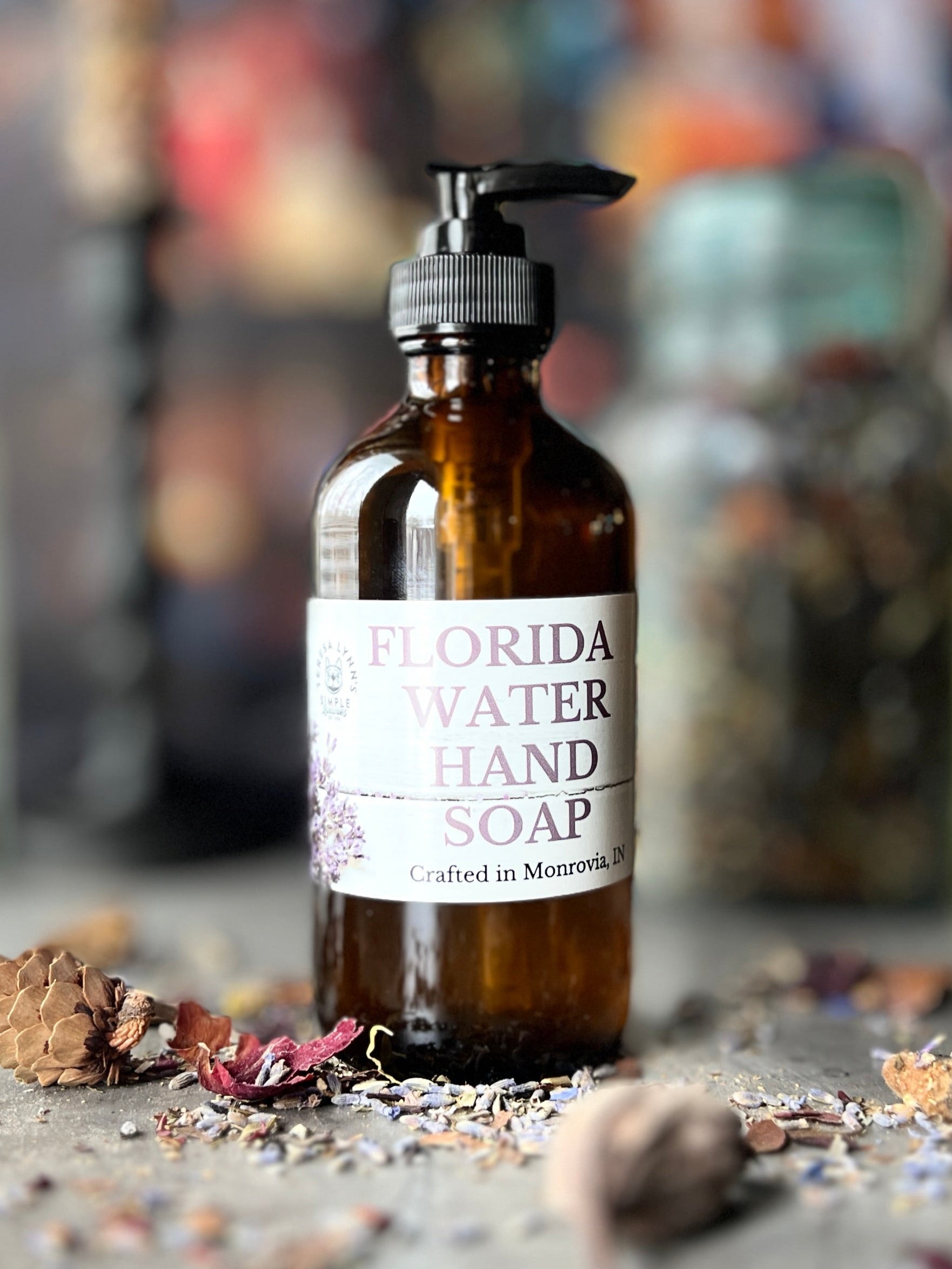 A brown glass bottle with a black pump top labeled Florida Water Hand Soap Crafted in Monrovia In sits on a grey surface with herbs and flowers scattered on it.  The background is a blurred collection of vintage bottles and jars.