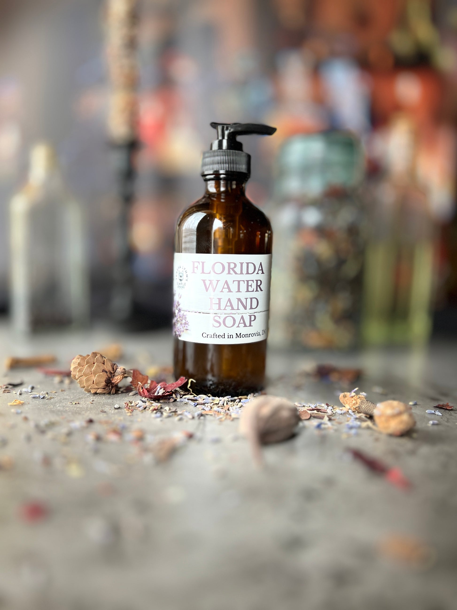 A brown glass bottle with a black pump top labeled Florida Water Hand Soap Crafted in Monrovia In sits on a grey surface with herbs and flowers scattered on it.  The background is a blurred collection of vintage bottles and jars.