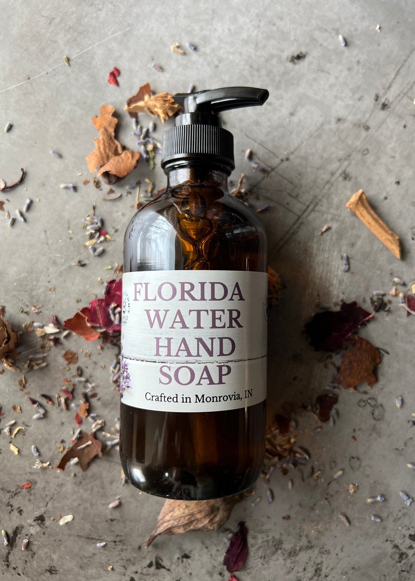 A brown glass bottle with a black pump top labeled Florida Water Hand Soap Crafted in Monrovia In sits on a grey surface with herbs and flowers scattered on it.  The background is a blurred collection of vintage bottles and jars.