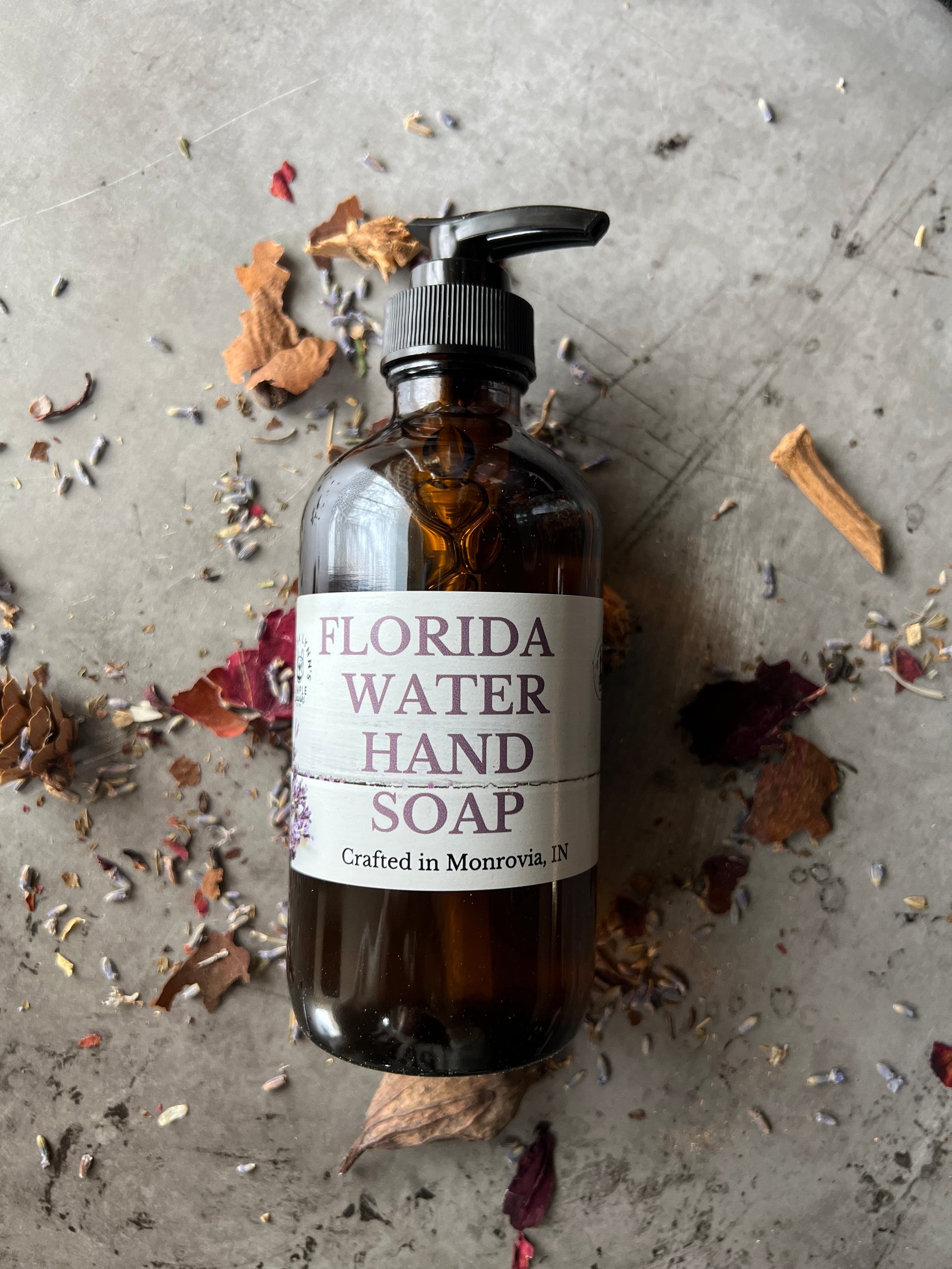 A brown glass bottle with a black pump top labeled Florida Water Hand Soap Crafted in Monrovia In sits on a grey surface with herbs and flowers scattered on it.  The background is a blurred collection of vintage bottles and jars.