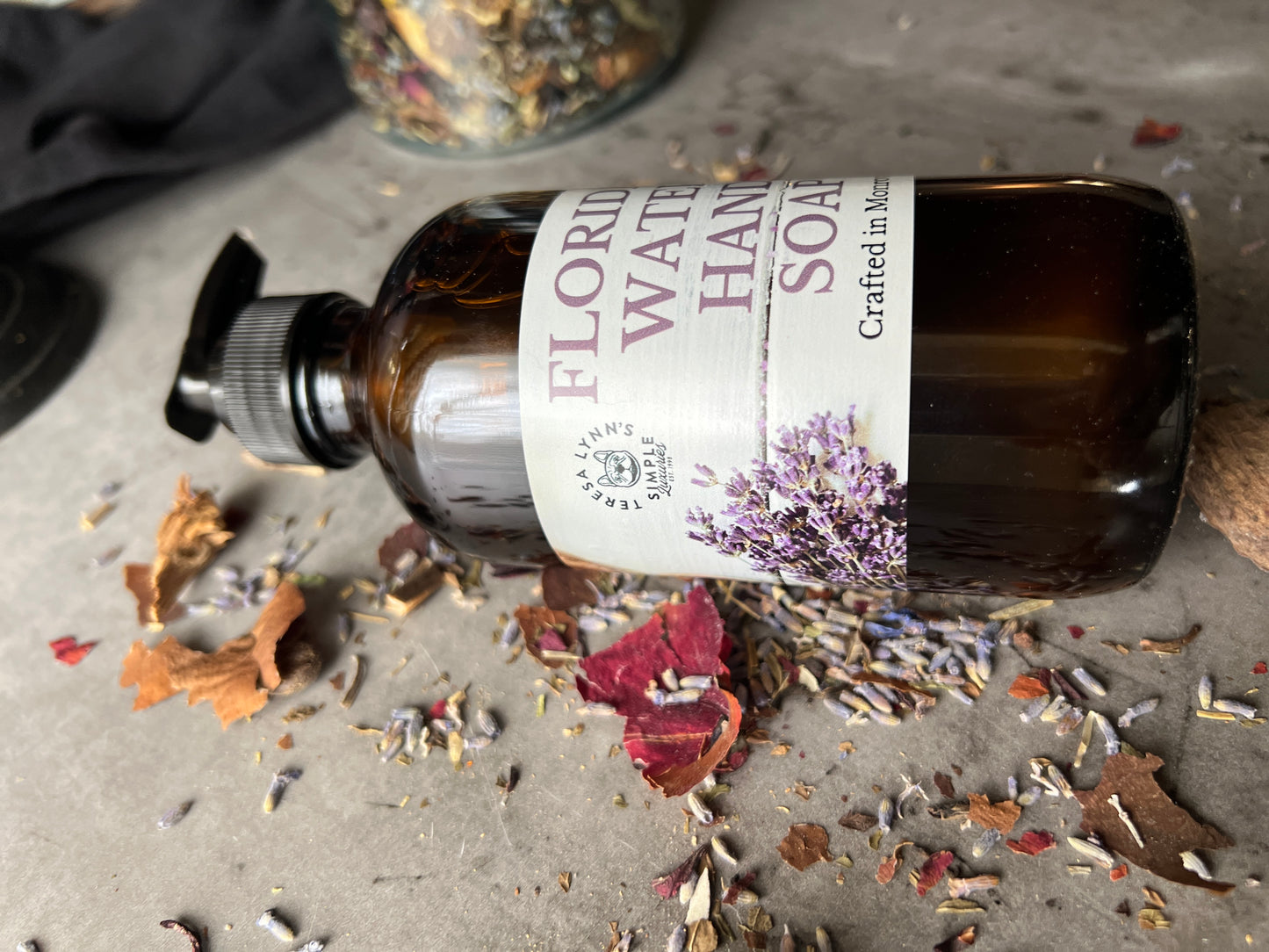 A brown glass bottle with a black pump top labeled Florida Water Hand Soap Crafted in Monrovia In sits on a grey surface with herbs and flowers scattered on it.  The background is a blurred collection of vintage bottles and jars.