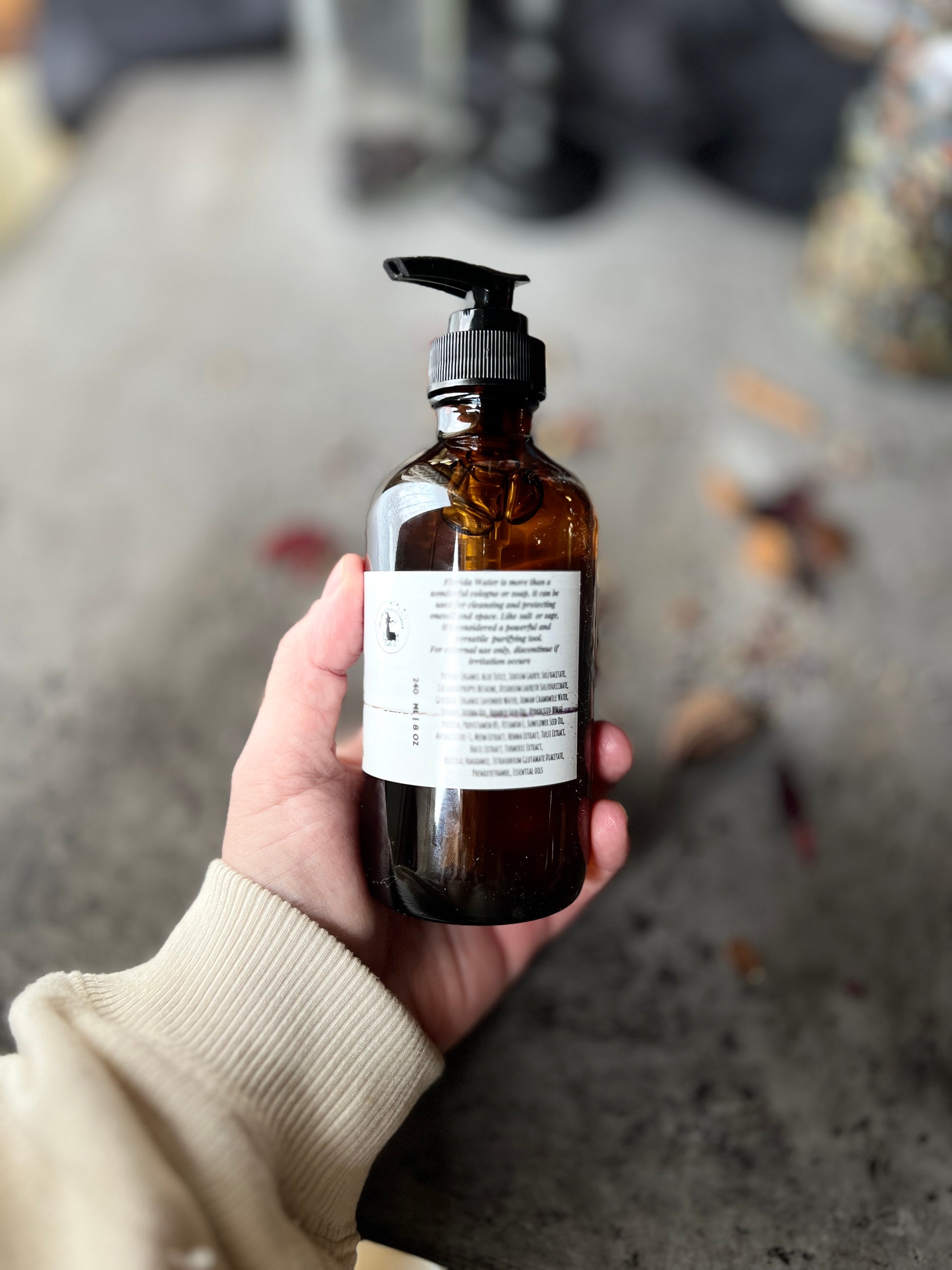 A brown glass bottle with a black pump top labeled Florida Water Hand Soap Crafted in Monrovia In sits on a grey surface with herbs and flowers scattered on it.  The background is a blurred collection of vintage bottles and jars.