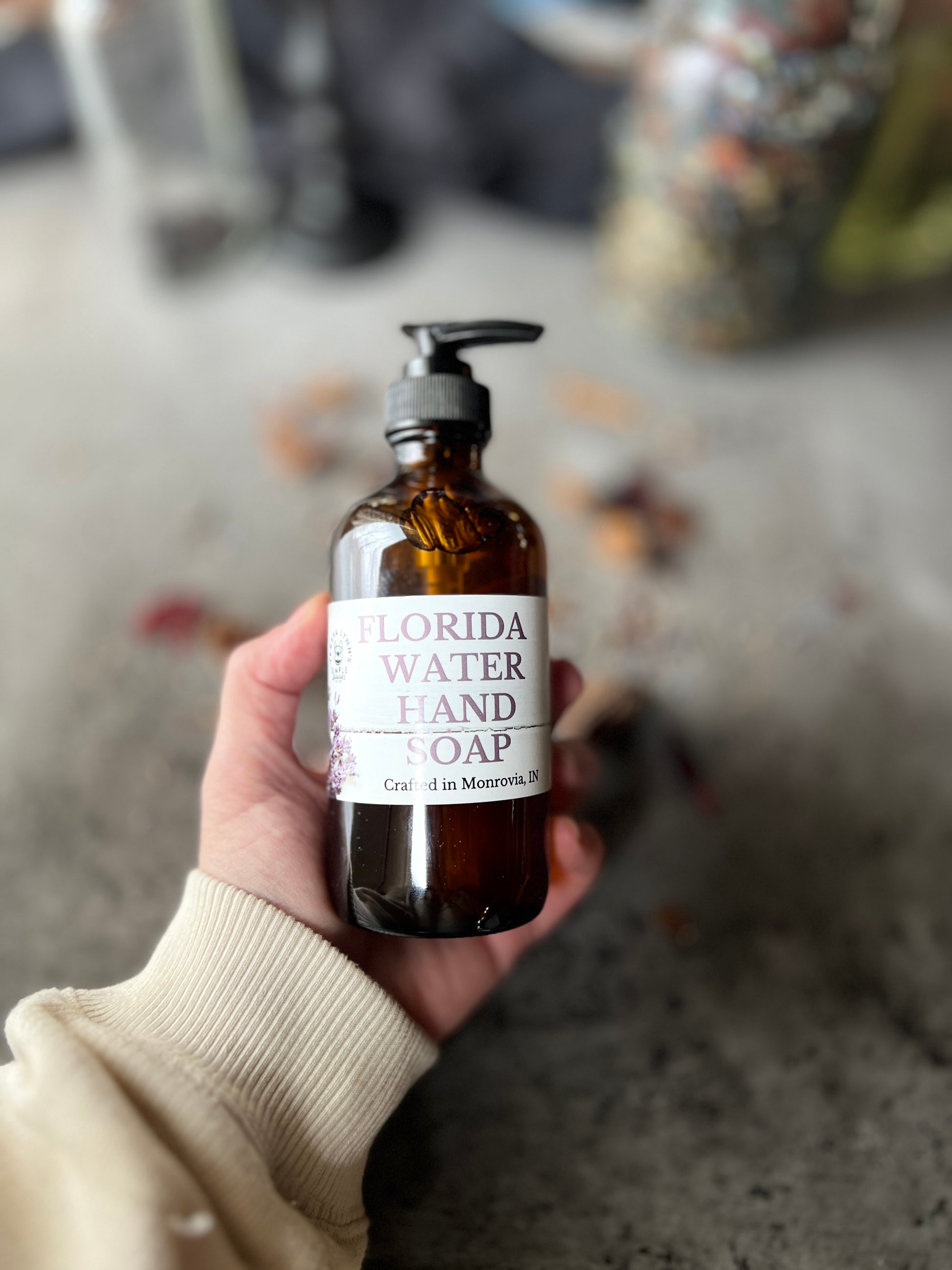 A brown glass bottle with a black pump top labeled Florida Water Hand Soap Crafted in Monrovia In sits on a grey surface with herbs and flowers scattered on it.  The background is a blurred collection of vintage bottles and jars.