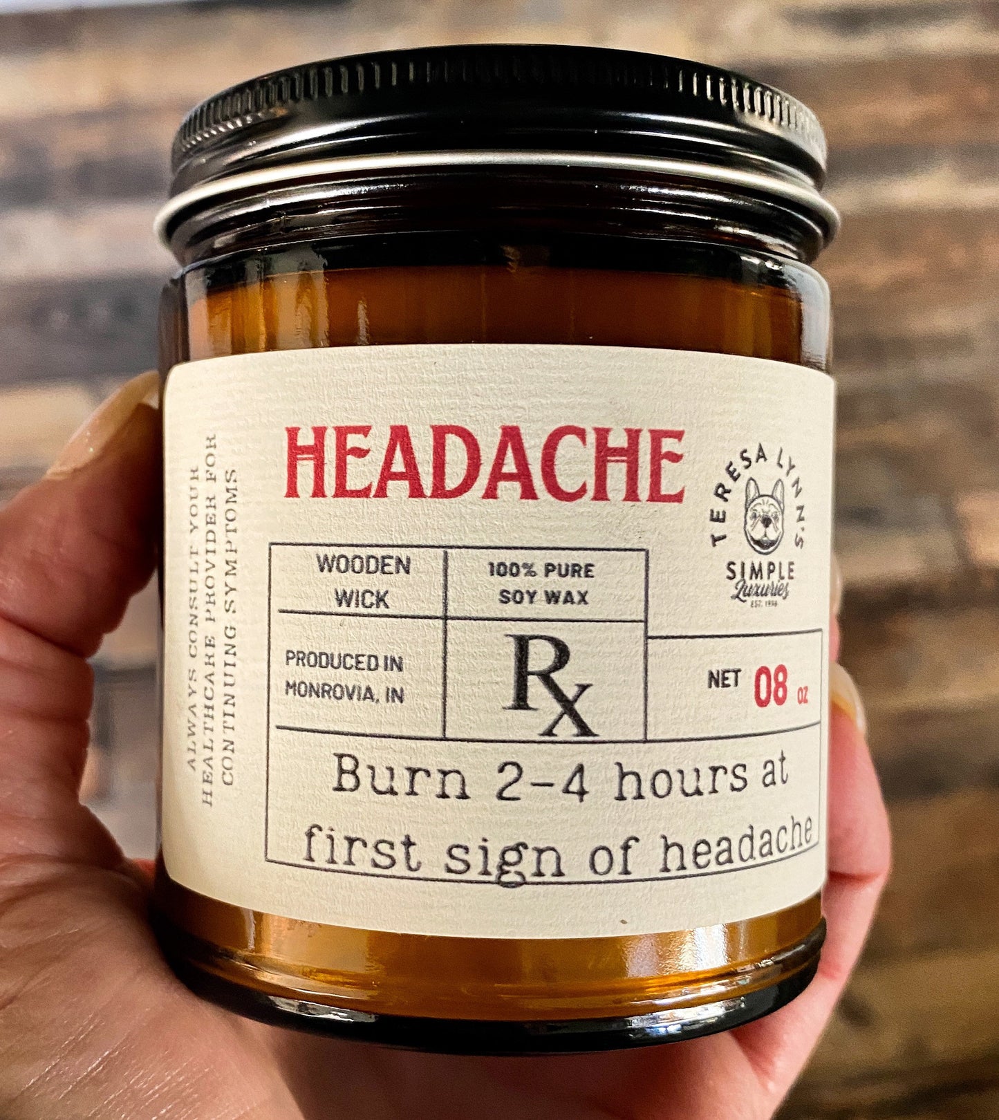 In Teresas Hand sits a 9oz brown glass apothecary style candle jar with a black lid.  The label is designed to look like a vintage prescription label with the word Headache at the top in red antique font.